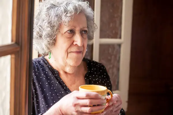 Seventy-year-old woman drinking a cup of coffee by a window while thinking about the things she can do during the day. A small smile appears on his face.
