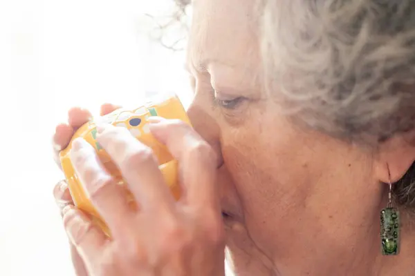Extreme close-up of seventy-year-old woman holding a cup of coffee in her hands as she sips it. The scene is illuminated by the daylight that enters through one of the windows of his house.
