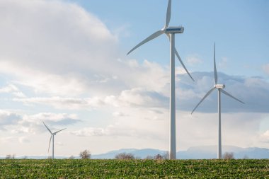 Üç rüzgar türbini bir tarlada duruyor, en uzun olanı ortada. Gökyüzü bulutlu, ama güneş hala görünür.