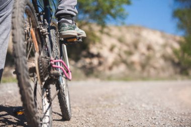 Bir insan ayakları pedalların üzerinde bisiklete biniyor. Bisiklet arka planda kayalık bir tepenin olduğu toprak bir yolda..
