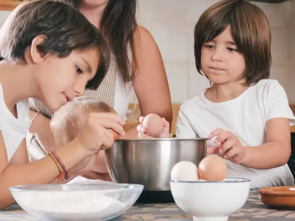 Üç çocuklu bir aile ve bir kadın birlikte kahvaltı hazırlıyorlar. Kadınlar izlerken çocuklar bir kaseye yumurta kırıyorlar. Sahne sıcak ve aile odaklı.