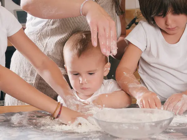 Bir kadın bir bebeğin pasta yapmasına yardım ediyor. Bebek una bulanmış ve yüzünde ciddi bir ifade var. Diğer çocuklar izliyor ve yardım etmek için sıralarını bekliyorlar. Sahne çok neşeli.