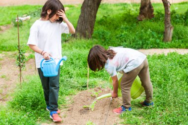 İki çocuk bahçedeki bitkileri suluyor. Bir tanesinin elinde su kovası var. Sahne huzurlu ve sakin, çocuklar dışarıda vakit geçiriyorlar.