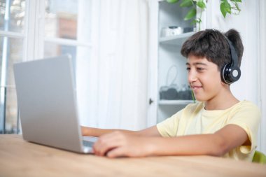 Bir çocuk dizüstü bilgisayarı ve kulaklığı olan bir masada oturuyor. Gülümsüyor ve eğleniyor.