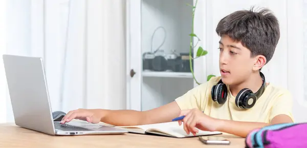 Bir çocuk dizüstü bilgisayarı ve not defteriyle masada oturuyor. Kulaklık takıyor ve işine odaklanmış durumda.