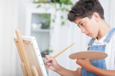 Kot pantolonlu çocuk bir fırça ve bir renk paleti ile resim bir tuval üzerine monte onun oturma odasında bir sehpa üzerinde. 