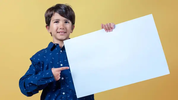 Bir çocuk beyaz bir poster tutuyor ve parmağıyla işaret ediyor. Poster beyazlar içinde. turuncu izole arkaplan. 