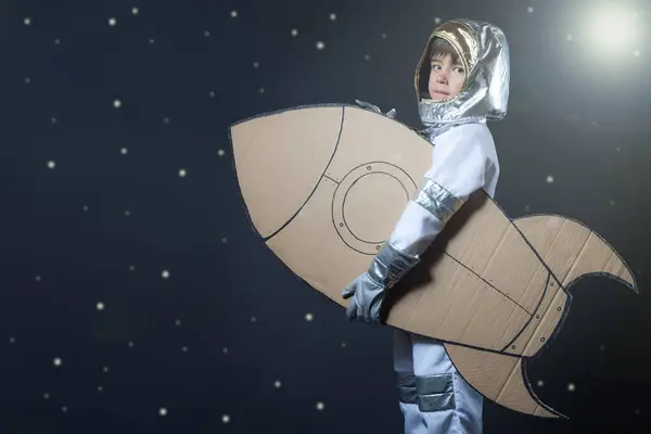 Astronot gibi giyinmiş bir çocuk yıldızlı arka planda karton bir roket tutuyor.. 