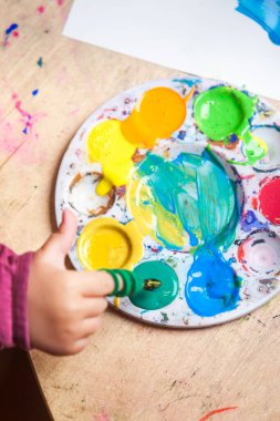 Akrilik renklerle dolu bir ressam paleti ve parmak fırçalı bir çocuk eli olan bir masanın ayrıntılı görüntüsü.. 