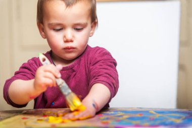 Yeni yürümeye başlayan çocuk eline altın boya sürüyor. Arkasında boş bir tuval var.. 
