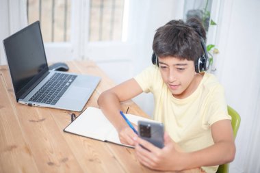 Bilgisayarlı ve not defterli bir masada çalışırken kulaklık takan bir genç cep telefonuyla çalışıyor..