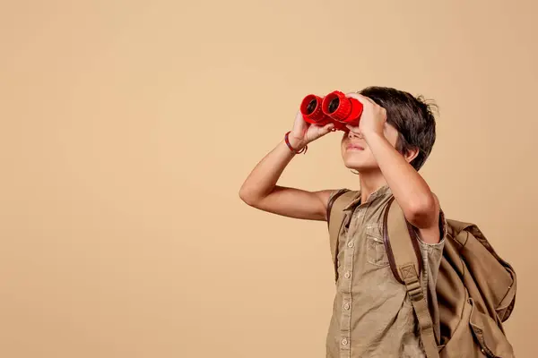 Sırt çantası taşıyan bir çocuk bronzlaşmış arka plandaki kırmızı dürbünle bakar. Merak ve macera kavramı
