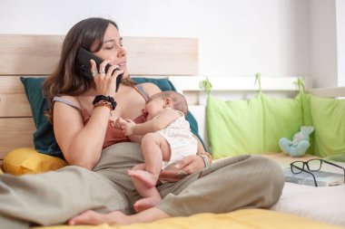 Bir kadın kucağında bir bebekle yatakta yatarken cep telefonuyla konuşuyor.. 
