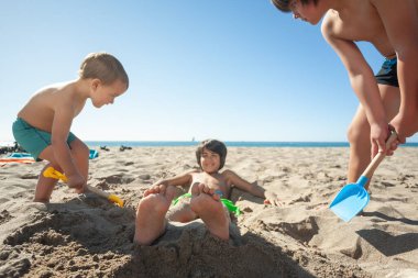 Üç çocuk kumsalda oynuyor. Bir çocuğu kuma kürekle gömmek için.. 