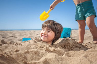 Gülümseyen bir çocuğun kafası kumdan fırlar ve arkasında da elinde kürekle tanınmayan bir çocuk.. 