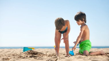Çocuklar güneşli bir kumsalda bir kürekle çukur kazıyor.