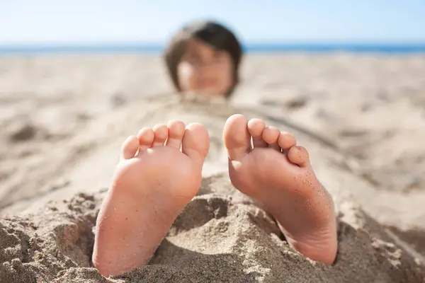 Sahildeki kumda gömülü bir çocuğun ayaklarının yakın çekimi. Arka planda kafası bulanık.. 