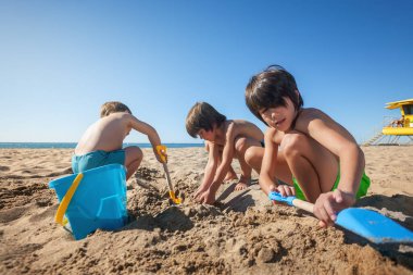 Üç çocuk kumsalda kürek ve kovayla oynuyor.. 