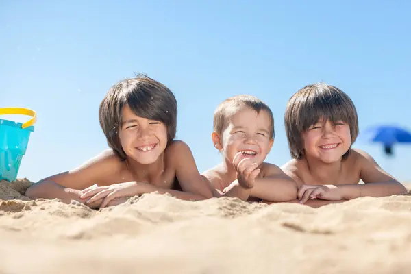 Üç çocuk kumsalda yatıyor, gülümsüyor ve güneşin tadını çıkarıyorlar.