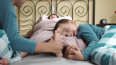 Bir çocuk yorganın içine sarılmış bir yatakta yatıyor ve küçük kardeşleri elleriyle ayaklarının tabanını gıdıklıyor. En küçüğü kardeşinin kucağında yatıyor..