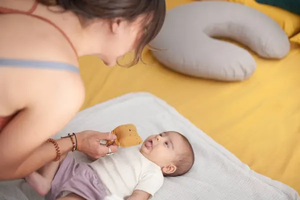 Bir kadın bebeğiyle yatakta oynuyor. Anne kızına yumuşak bir oyuncak gösterir..