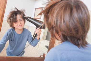 Mavi gömlekli genç bir çocuk, dağınık saçını saç kurutma makinesiyle kurutur. Aynaya bakar, bir ev banyosunda neşeyle gülümser..