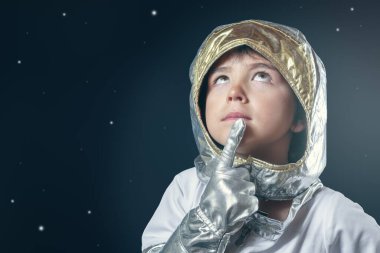 Uzay giysisi giymiş bir çocuk yıldızlara dalgın dalgın bakıyor. Eldiven ve kask takıyor.