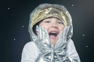 Gümüş rengi uzay elbisesi giymiş genç bir çocuk gülümsüyor ve komik bir surat yapıyor. Resmin neşeli ve neşeli bir havası var, çünkü çocuk astronot gibi giyinmiş ve eğleniyor.