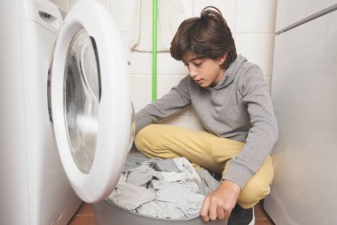 Bir çocuk bir çamaşır makinesinin önünde yerde oturuyor. Bir yığın giysiyi ayırıyor.