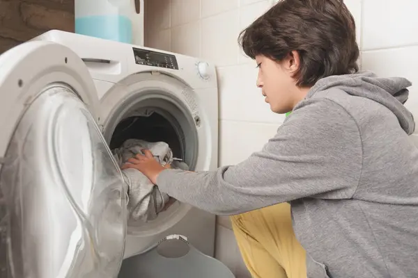 Bir çocuk kirli çamaşırları çamaşır makinesine koyar. Gri bir kapüşonlu giyiyor.