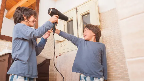 İki genç çocuk banyoda saç kurutma makinesiyle oynuyorlar. Çocuklardan biri kurutucuyu tutarken diğeri de arkasında duruyor. İkisi de gülümsüyor ve eğleniyor gibi görünüyorlar.