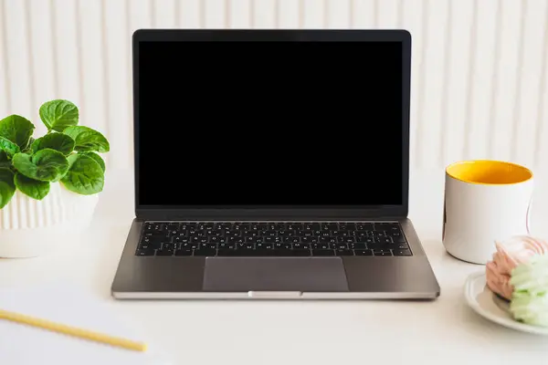 Mock-up digital laptop with empty screen, notebook, coffee cup, food and plant on white table. Front view