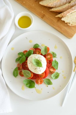 Beyaz ahşap masada domates, kiraz, mozzarella ve fesleğen yapraklı Caprese salatası.