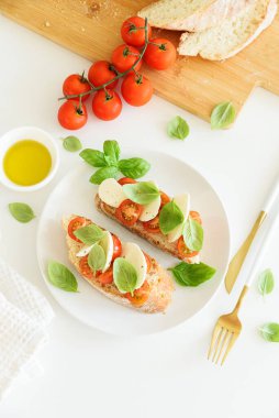 Beyaz ahşap masada mozzarella, kiraz domatesi ve fesleğen yapraklı kızarmış ekmek.