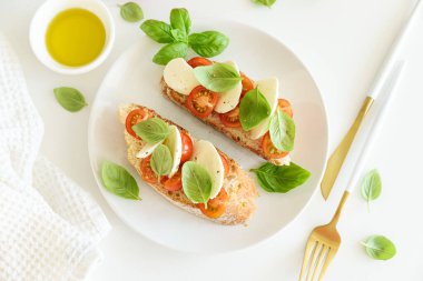 Beyaz ahşap masada mozzarella, kiraz domatesi ve fesleğen yapraklı kızarmış ekmek.