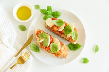 Beyaz ahşap masada mozzarella, kiraz domatesi ve fesleğen yapraklı kızarmış ekmek.