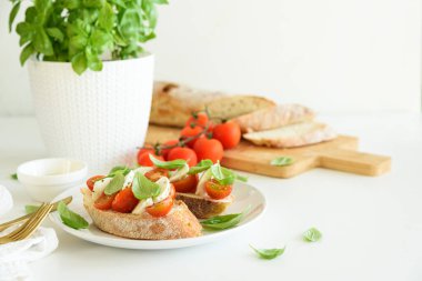 Beyaz ahşap masada mozzarella, kiraz domatesi ve fesleğen yapraklı kızarmış ekmek.