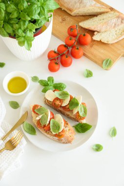 Beyaz ahşap masada mozzarella, kiraz domatesi ve fesleğen yapraklı kızarmış ekmek.
