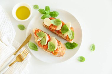 Beyaz ahşap masada mozzarella, kiraz domatesi ve fesleğen yapraklı kızarmış ekmek.