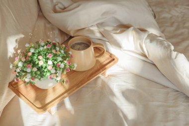 Yatakta kahvaltı: Tahta tepside bir fincan kahve ve buket. Pencere ışığı