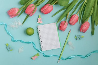 Pastel Paskalya sahnesinde mavi bir kurdele, bir defter, ahşap tavşan heykelcikleri ve yumuşak tüylerle süslenmiş pembe laleler var. Paskalya tatilini kutlamak için mükemmel..