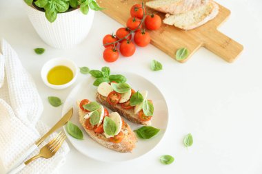 Beyaz ahşap masada mozzarella, kiraz domatesi ve fesleğen yapraklı kızarmış ekmek.