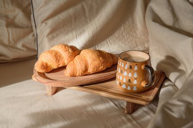 Yatakta kahvaltı: bir fincan kahve ve ahşap tepside taze kruvasan. Sıcak pencere ışığı