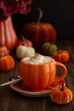 Balkabağı şeklinde bir kupada, sonbahar dekoruyla çevrili, krema ve baharatlı, kabak şeklinde bir latte.