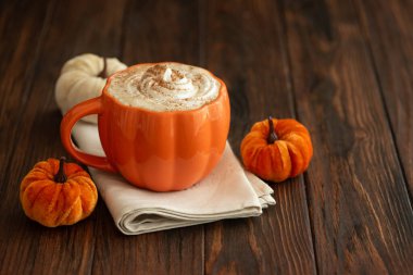 Balkabağı şeklinde bir kupada, sonbahar dekoruyla çevrili, krema ve baharatlı, kabak şeklinde bir latte.