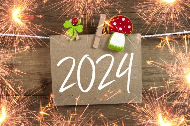Wooden hang tag and slate with four leaf clover and sparklers with the german words for happy new year - frohes neues jahr 2024 on wooden weathered background clipart