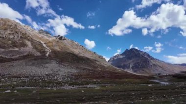 4K Nehir Manzarası Önden Arkaya Hareketli Kamera Görüntüsü Taşlar ve Çimenlerle Akan Nehir, Görkemli Rocky Dağları Tepeleri, Güzel Mavi Gökyüzü Fluffy ve Dinamik Beyaz Bulutlar