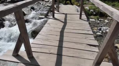 Güneşli bir günde, Rocky Sahilleri ile Taşlar Arasındaki Hızlı Akan Kristal Berrak Dağ Nehrinin Üzerinde Tahta Köprüde Yürüyorum