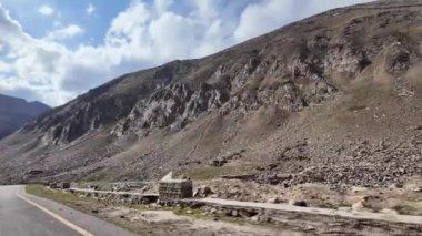 Yeşil Çayırlarda Dolaşan Kara Bitumen Yolu 'nun yan görüntüsü Çimen ve Taşlı Görkemli Dağ Sırası tarafından çerçevelenmiş. Yukarıda, güzel mavi gökyüzü beyaz kabarık bulutlarla süslenir, güneş ışığı