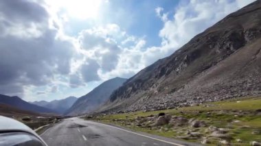 Green Meadows 'un yanındaki Kara Bitumen Yolu' nun yan görüntüsünde İneklerin yemyeşil bir dağ sırasının ortasında otladığı yerler taşlarla süslenir. Yukarıda, bir Mavi Gökyüzü Yumuşak Beyaz Bulutlarla dolu.